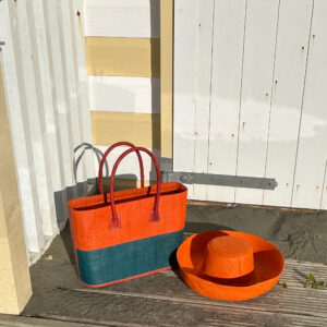 Monique raffia handbag with zip fastening in orange and turquoise with orange Mimosa raffia hat beside a beach hut